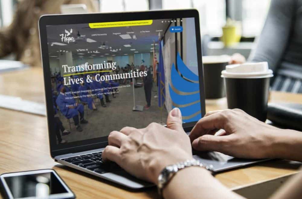 Hands type on a laptop displaying a "Hope" website with "Transforming Lives & Communities" text and people in blue.