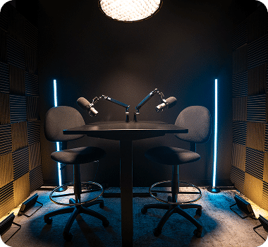 A dark soundproofed podcast studio with two microphones, two chairs, and blue and yellow LED lights.