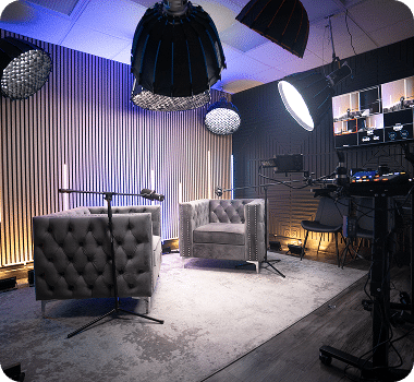 A modern podcast studio with two gray tufted armchairs, microphones, and professional lighting equipment.