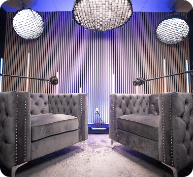 Two gray tufted armchairs face a light wood slat wall, illuminated by blue and purple lights and three large circular lamps.