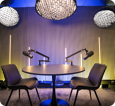 A modern podcast studio with two microphones, two chairs, a round table, and acoustic paneling.
