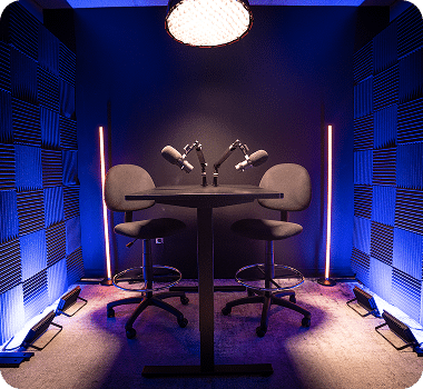 A small, dark podcast studio with two microphones, two chairs, and soundproofing on the walls.