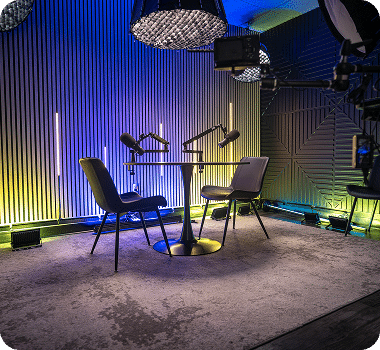 A modern podcast studio with two microphones on a round table, two chairs, and blue and yellow lighting.