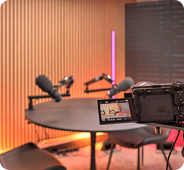 A camera on a tripod faces a round table with two microphones in a podcast studio with orange lighting.