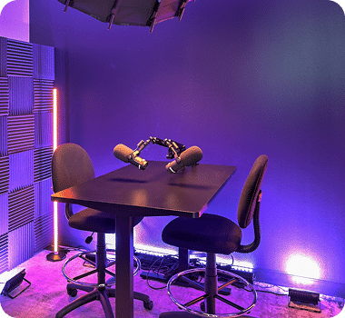A small podcast studio with purple lighting, a black table, two chairs, and two microphones.