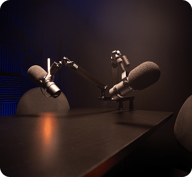 Two black microphones on adjustable arms sit on a dark table in a recording studio with blue acoustic panels.