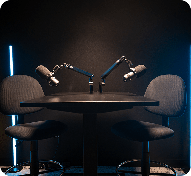 Two microphones on boom arms face each other across a dark table with two chairs in a dim room with blue neon light.