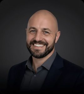 A smiling bald man with a dark beard and blue eyes wears a dark gray shirt and a navy suit jacket.