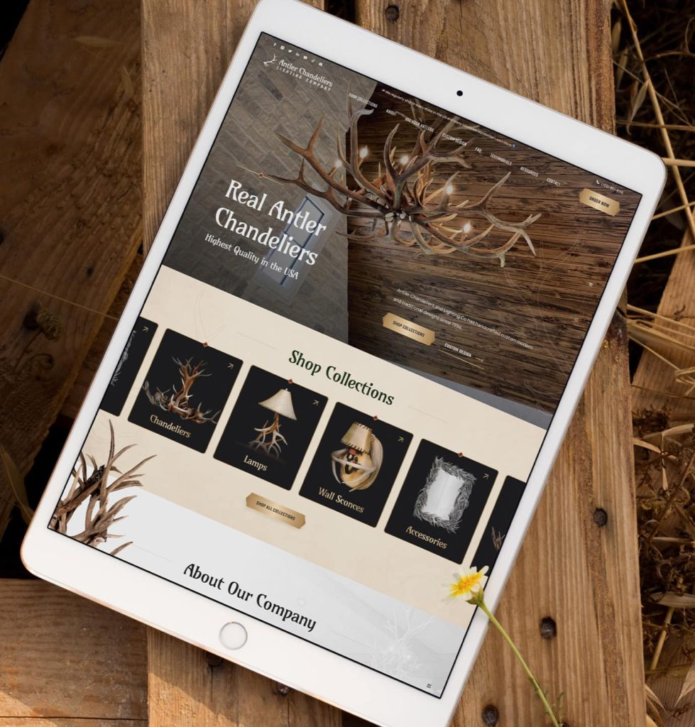 A white tablet displays "Antler Chandeliers Lighting Company" website on a rustic wooden surface with dried grass.