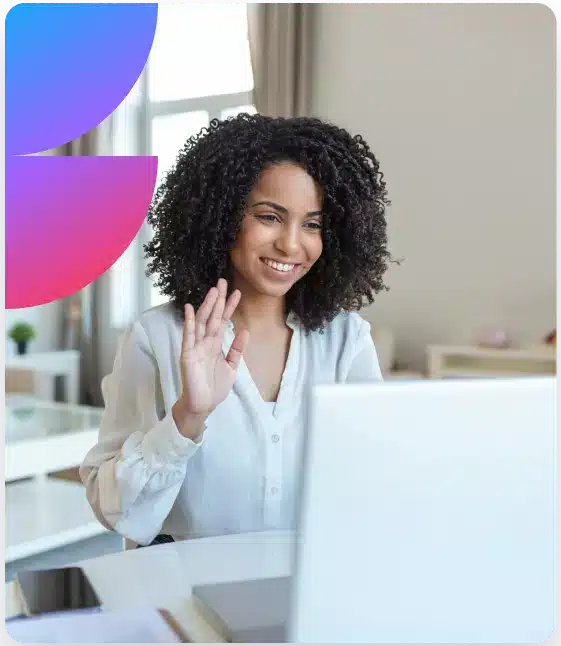A smiling woman with curly hair waves at a laptop while sitting at a desk with a phone and papers.