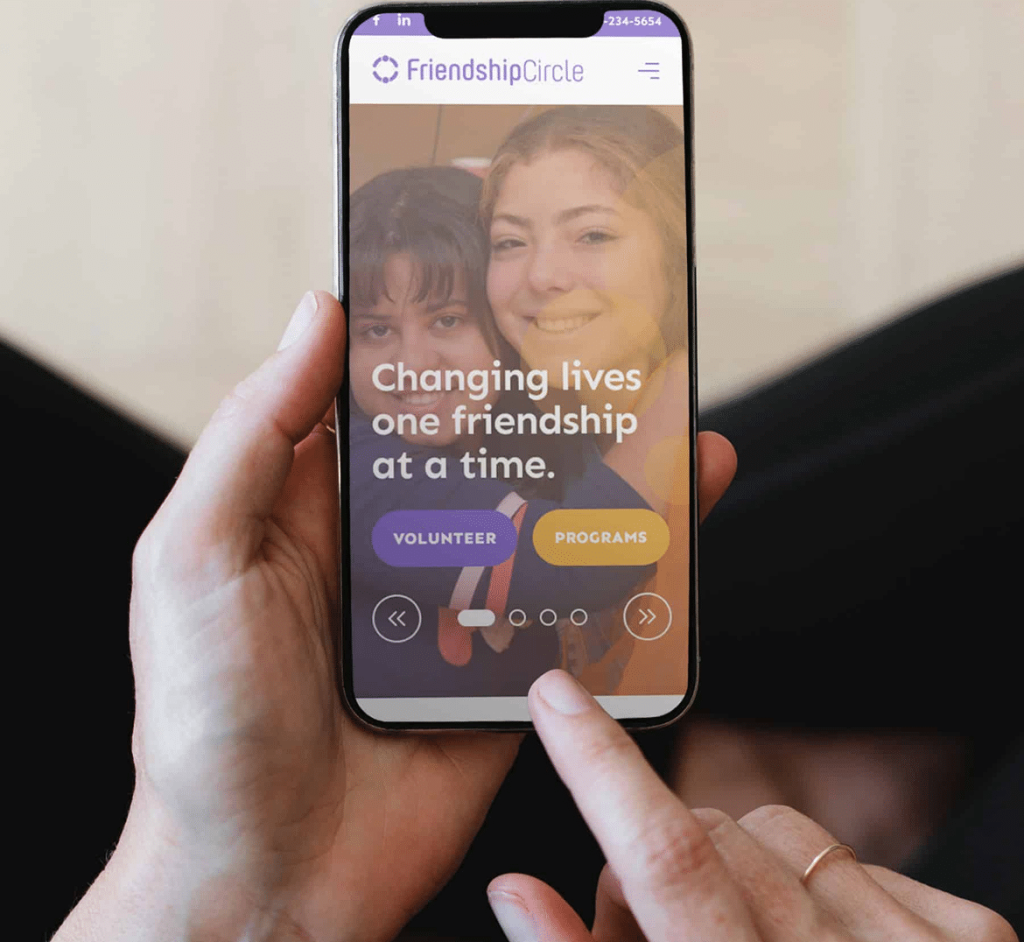 Hands hold a phone displaying the "FriendshipCircle" app with two smiling girls and text: "Changing lives one friendship at a time."