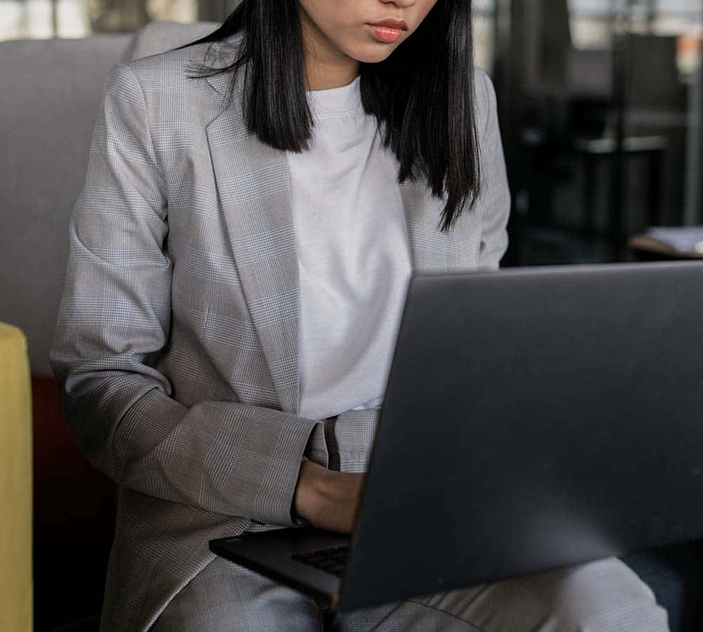 A young woman in a gray plaid blazer and white shirt sits, looking down at a black laptop.