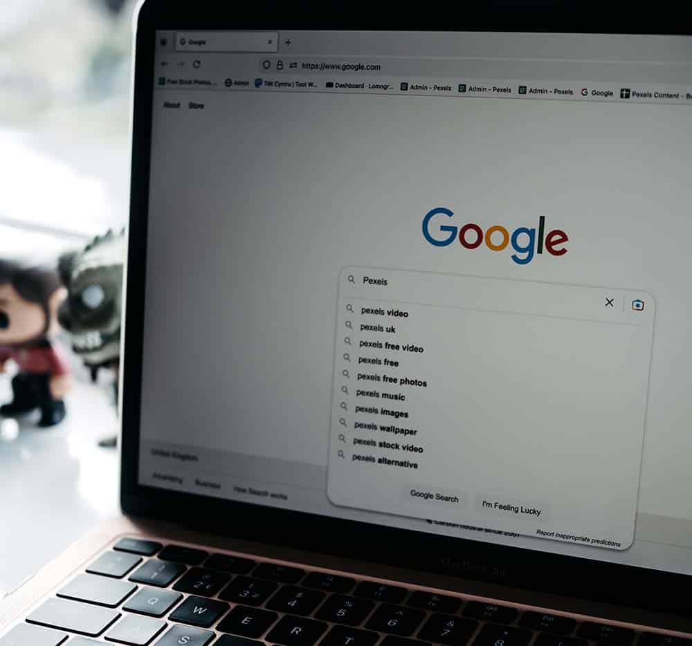 A laptop screen displays the Google search page with "Pexels" typed in the search bar and suggested results.