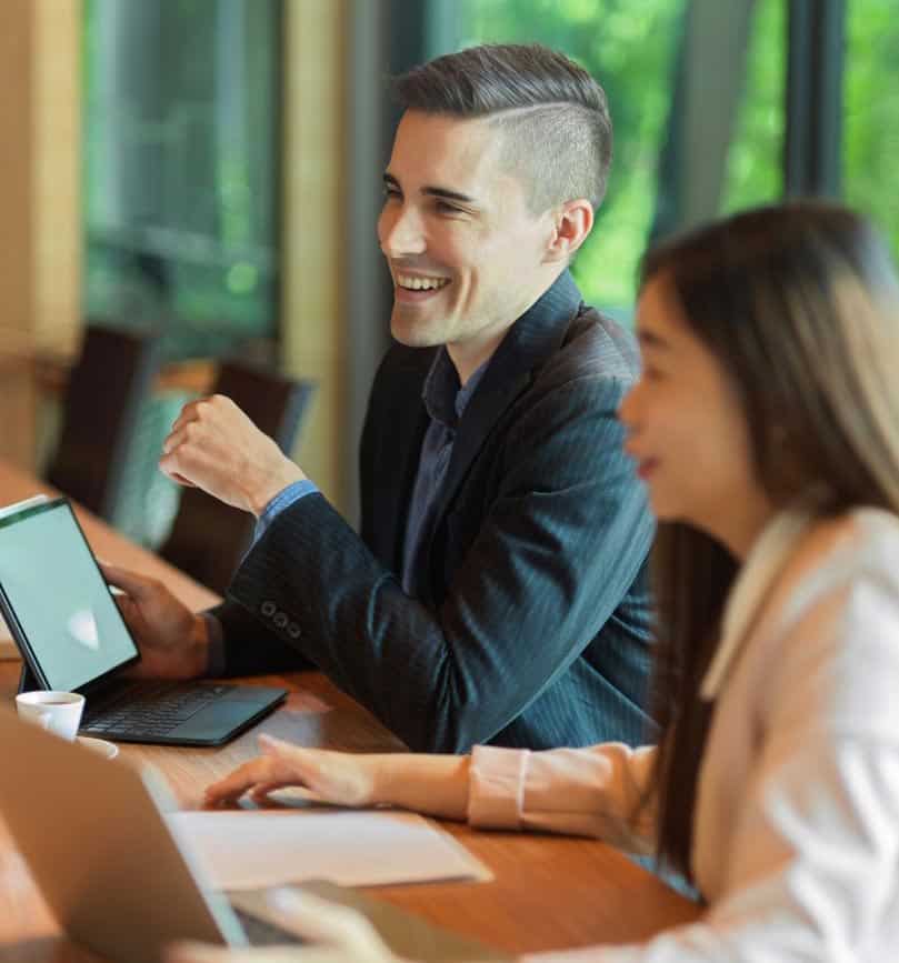 A smiling man in a suit holds a tablet with a white screen, sitting next to a woman at a table.