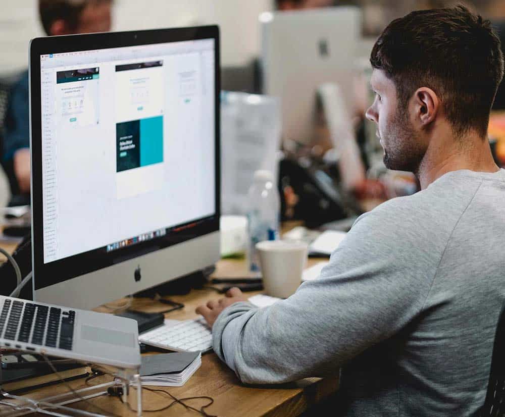 A man in a gray shirt works at a desk, looking at a computer screen displaying a website design.