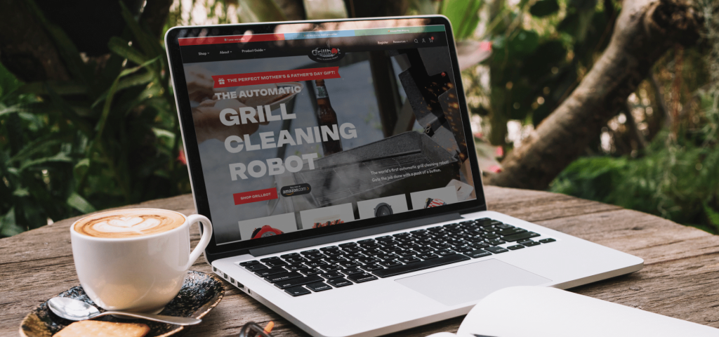 A laptop displaying "THE AUTOMATIC GRILL CLEANING ROBOT" website, with a latte and cookie on a wooden table.