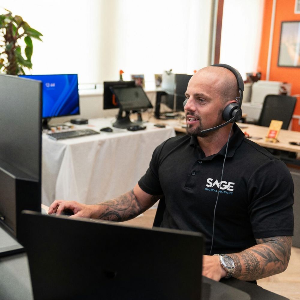 A man with a headset and "SAGE DIGITAL AGENCY" shirt works at a computer in an office.