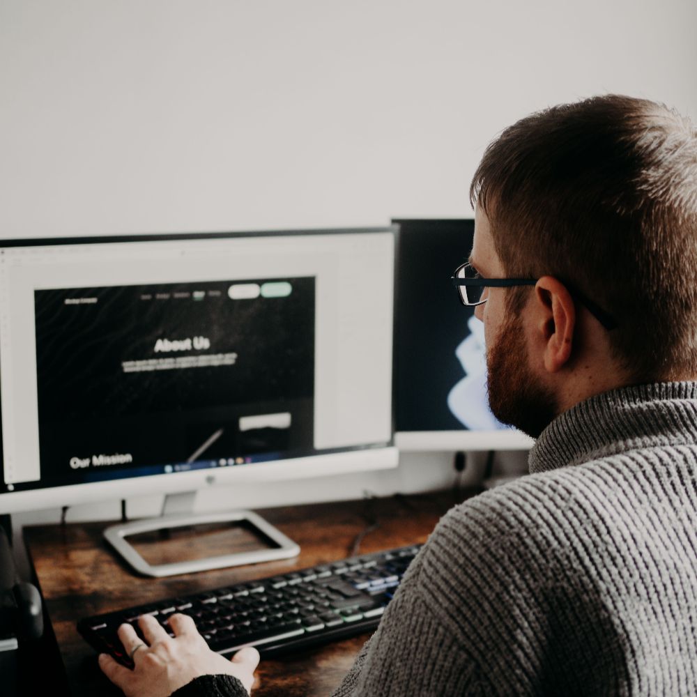A man with glasses and a beard works at a computer, viewing a webpage titled "About Us" and "Our Mission."